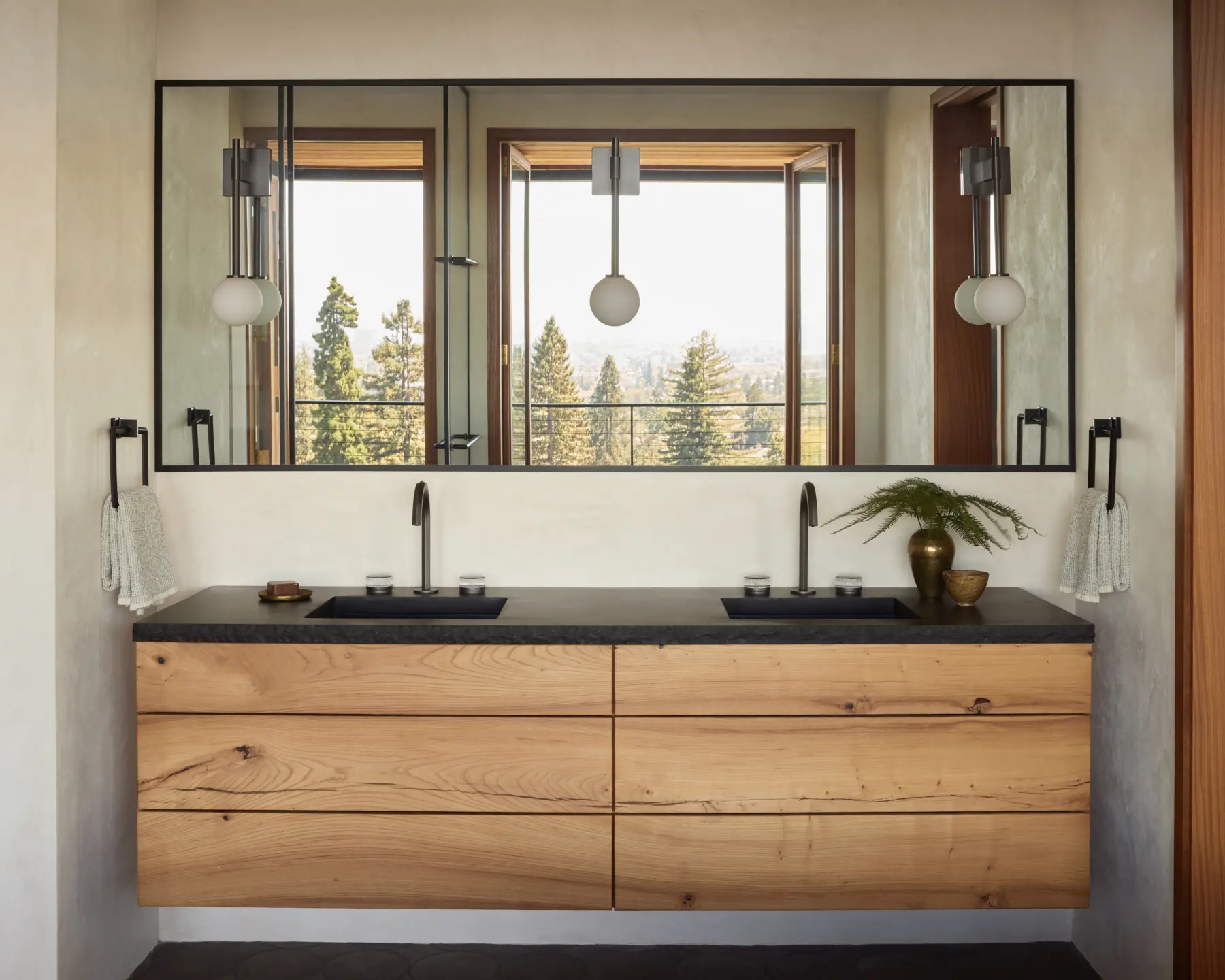 Interior bathroom with floating double wooden vanity, black countertop, two sinks, wall-mounted taps; black-framed mirror; hillside trees outside.