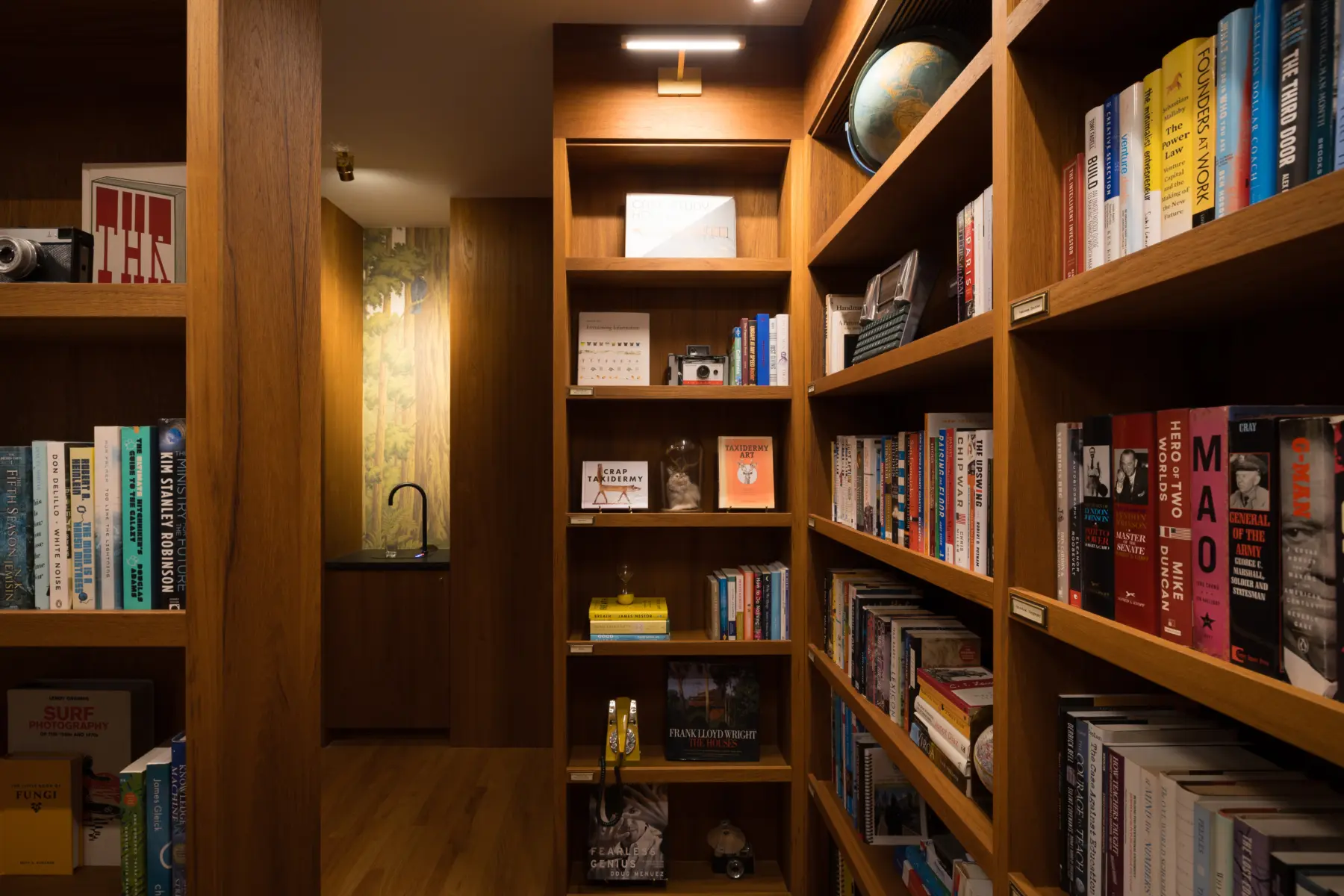 Interior view of warm oak library with tall built-in bookshelves on three walls, globe, and small black sink alcove; ceiling light; wood flooring.