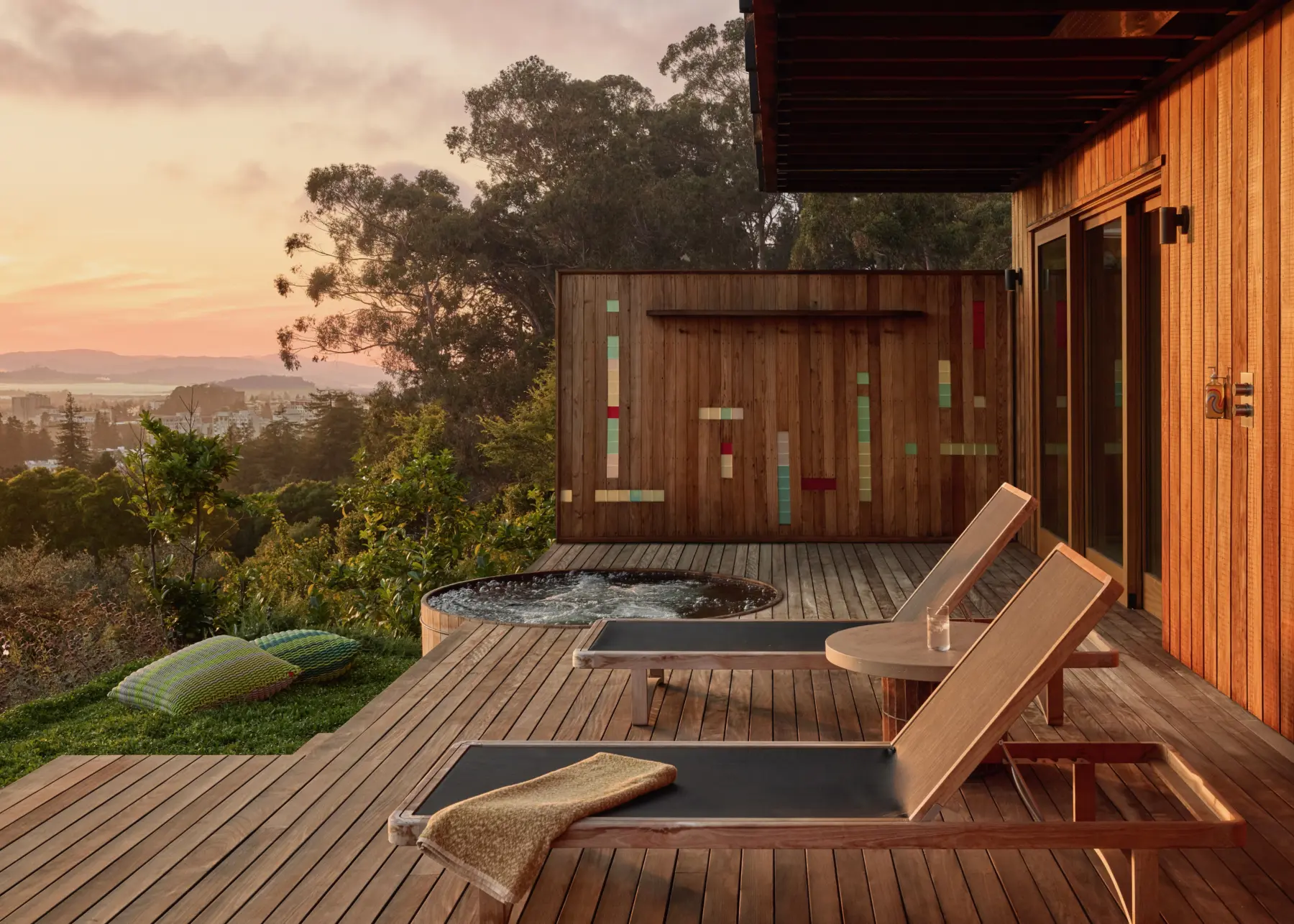 Exterior wood-clad hillside residence deck at sunset; round spa, two lounge chairs, sliding doors, and a colorful screen with glass strips; view.