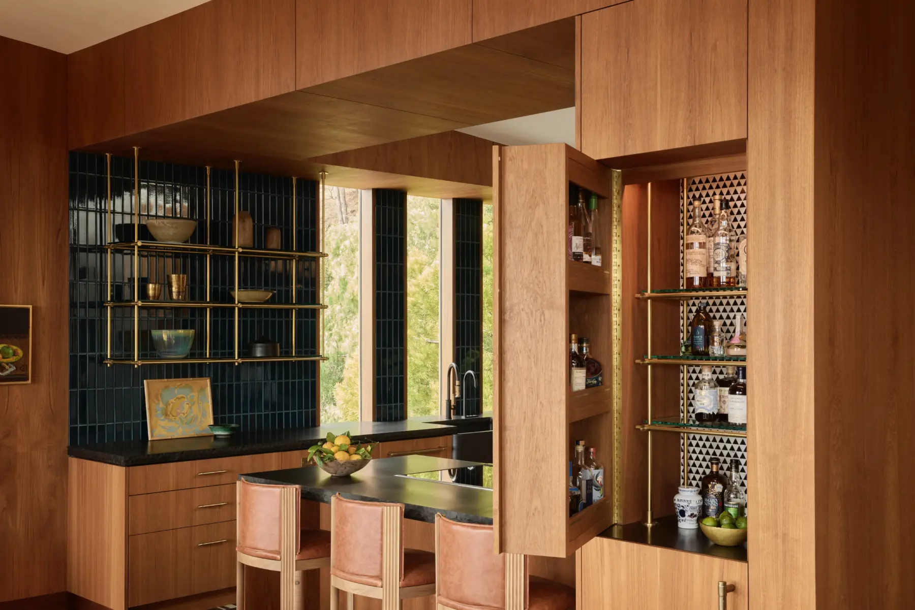 Interior: walnut-wood kitchen/bar with brass open shelves, dark blue tile backsplash, black countertop, and pink leather bar stools.