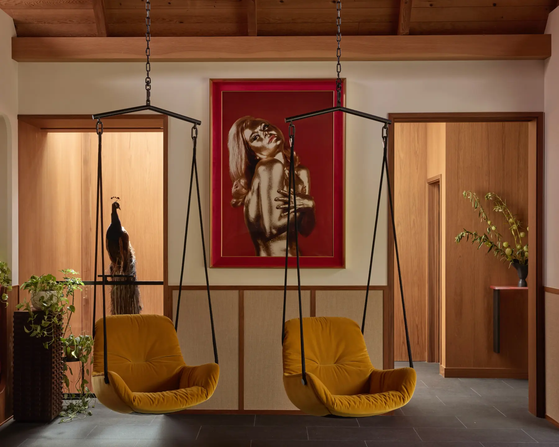 Interior foyer with timber beams; two mustard-yellow hanging chairs on black chains; red-framed painting of a woman; wood-paneled walls.