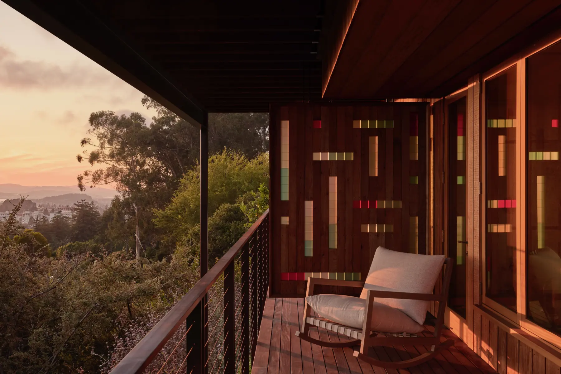 Exterior hillside balcony; dark wood siding with vertical colorful glass panels; deck, metal railing, cushioned chair at sunset, trees.