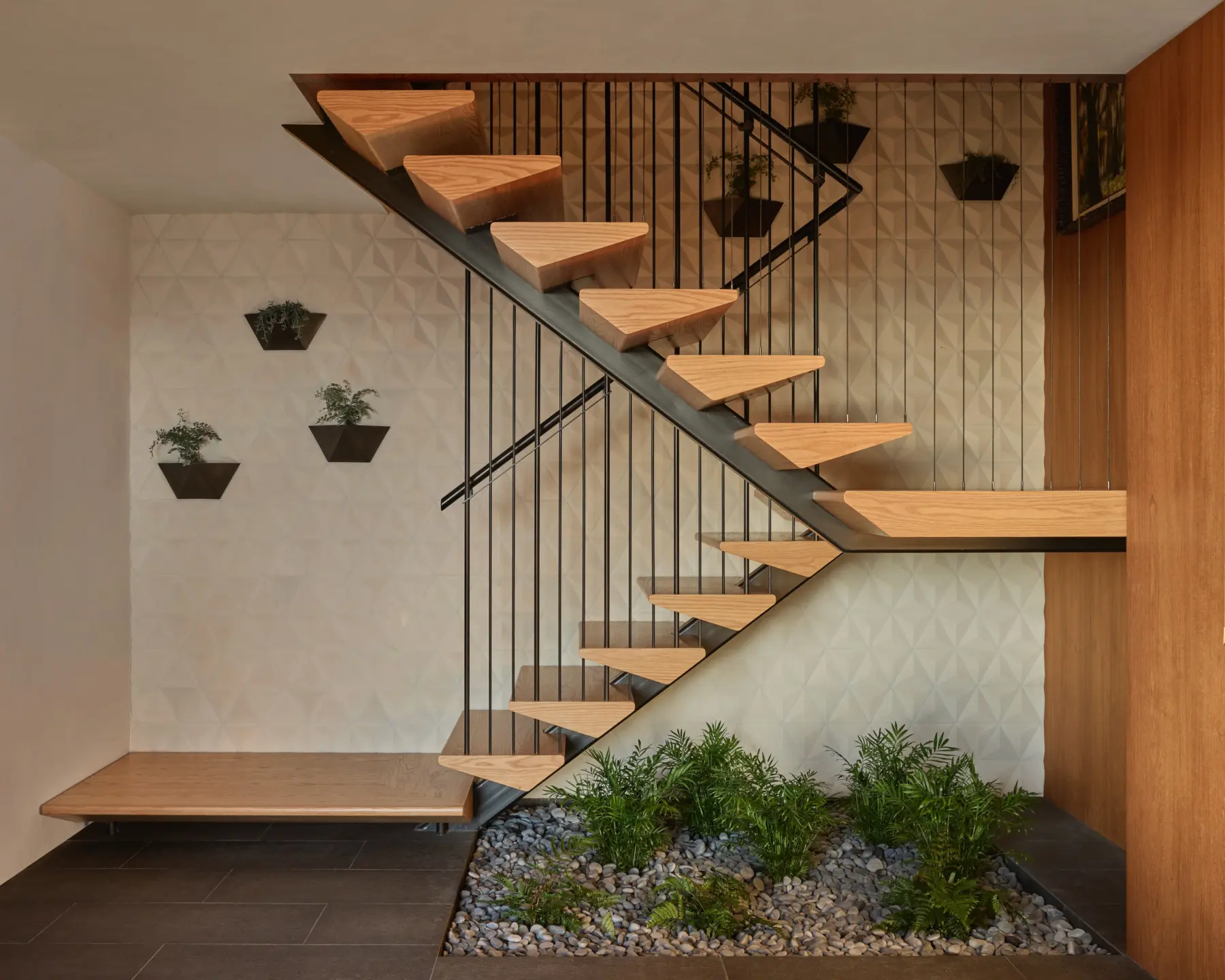 Interior staircase with open-riser wooden treads and black metal railing, alongside geometric-textured wall and indoor pebble garden.