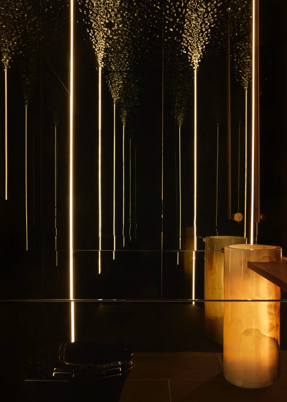 Moody interior with black wall panels and LED light columns; gold-toned marble cylinders beside a counter, creating a sculptural space.