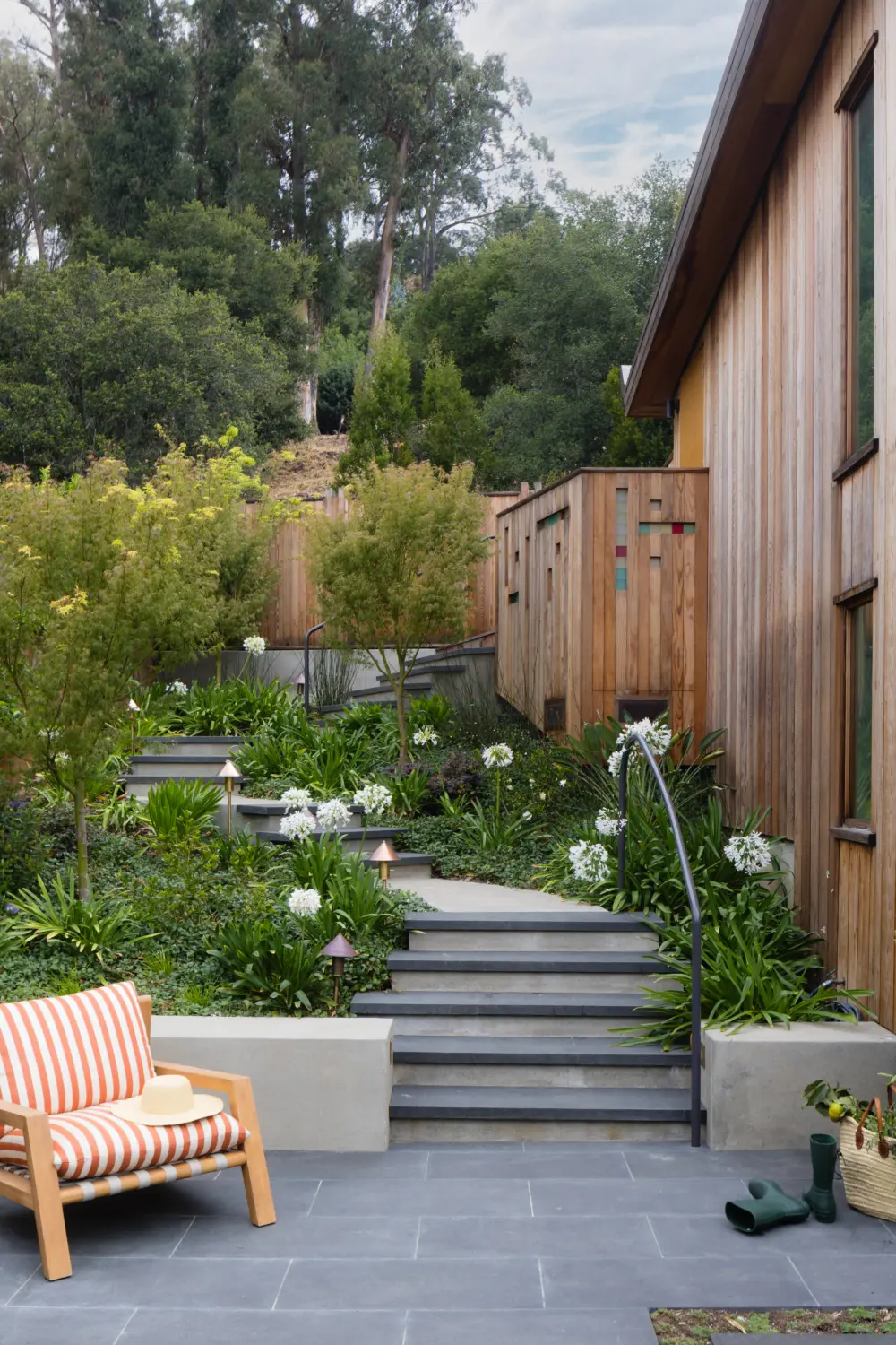 Wood-clad residence with tiered concrete steps and black metal handrail, lush drought-tolerant garden, slate-tiled patio, and striped-cushion chair.