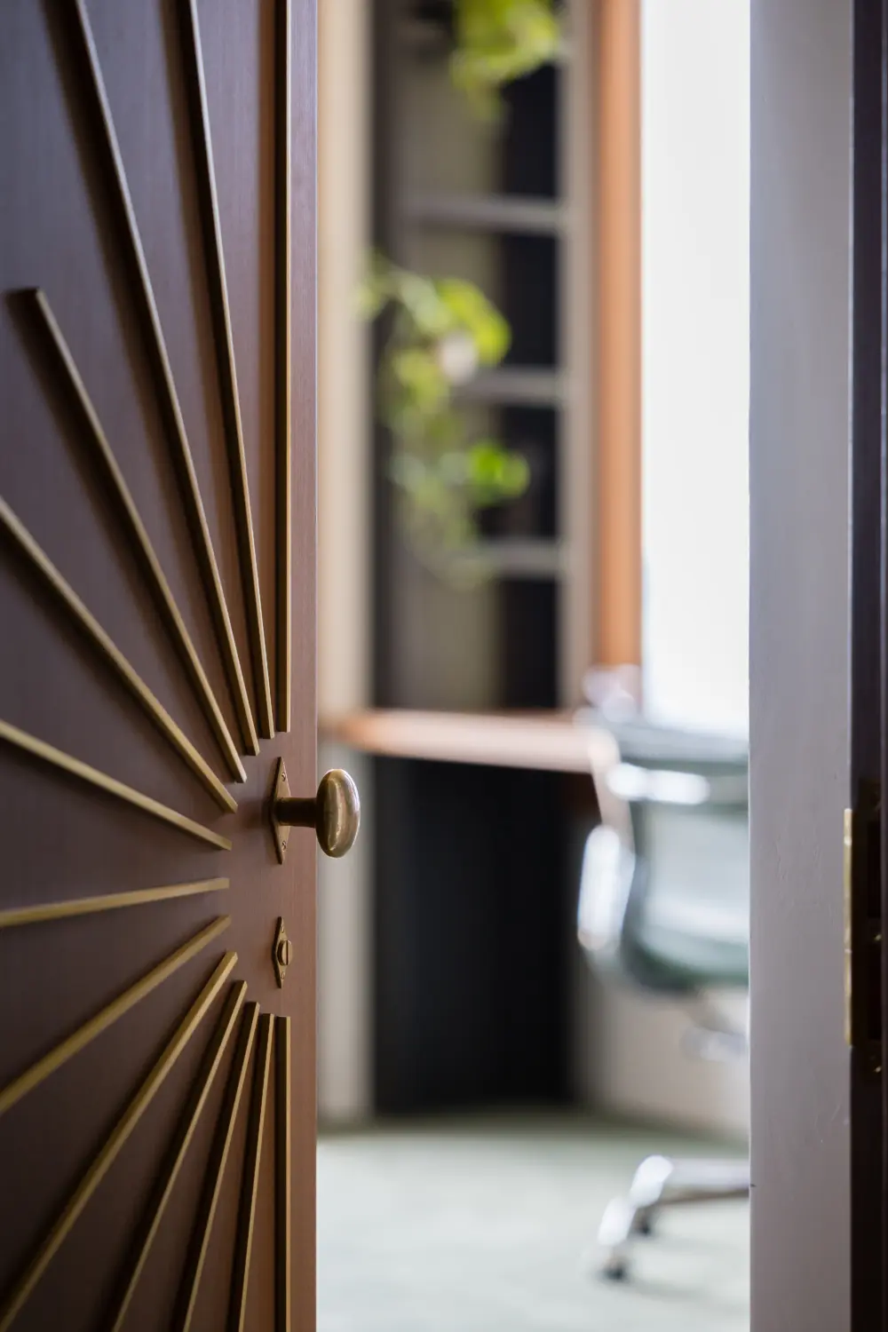 Wooden door with radial sunburst pattern and brass knob opens to a modern interior with a swivel chair, blurred desk and greenery.