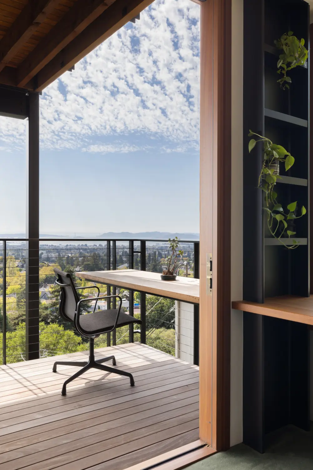 Balcony workspace on modern hillside residence: wooden desk, black office chair, potted plant, wood door frame, black metal railing, cityscape view.