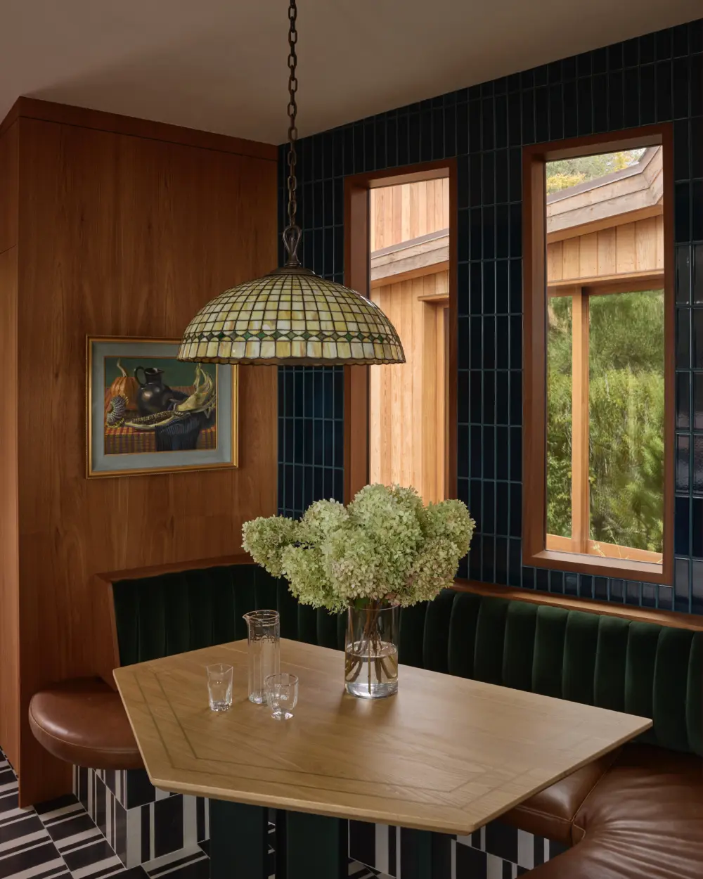 Dining nook with light wood table, hydrangea vase, dark green banquette, blue tile walls, Tiffany pendant lamp, wood-framed windows.