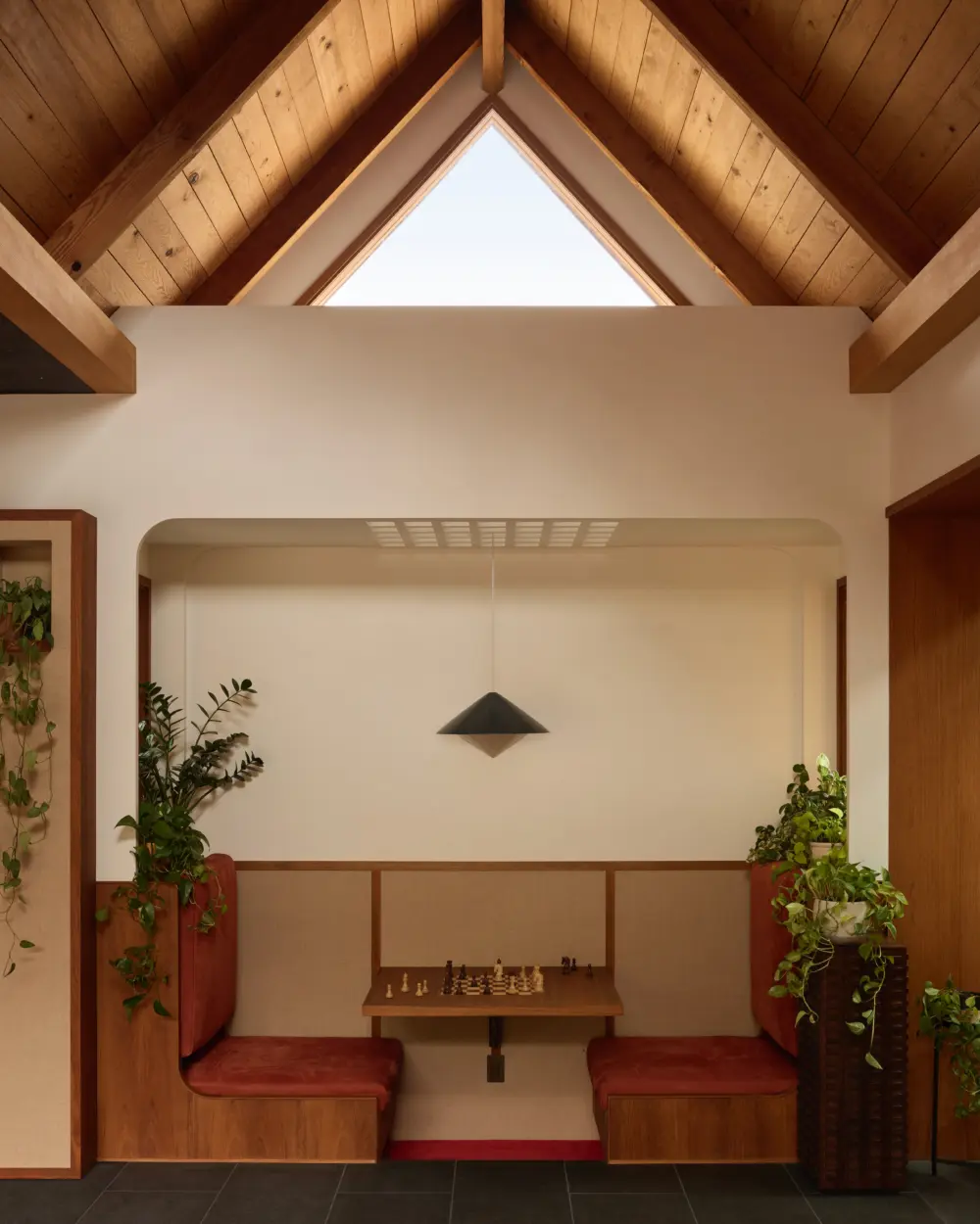 Interior dining booth under vaulted timber ceiling with triangular skylight; walnut banquettes, small chess table, black pendant, and potted plants.
