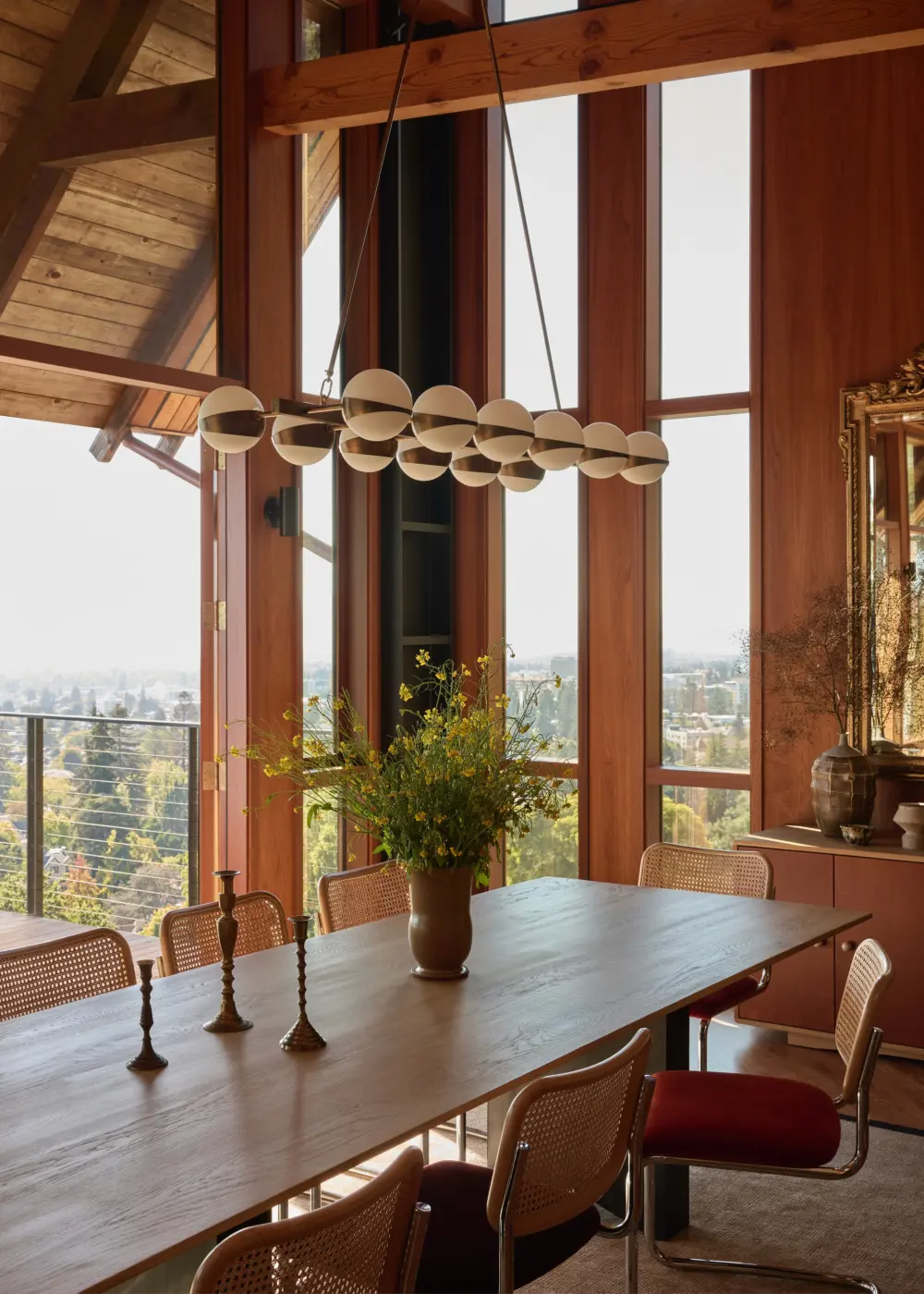 Dining room with long wood table, cane-back chairs, globe pendant chandelier, exposed timber beams, walnut windows framing hillside city view.