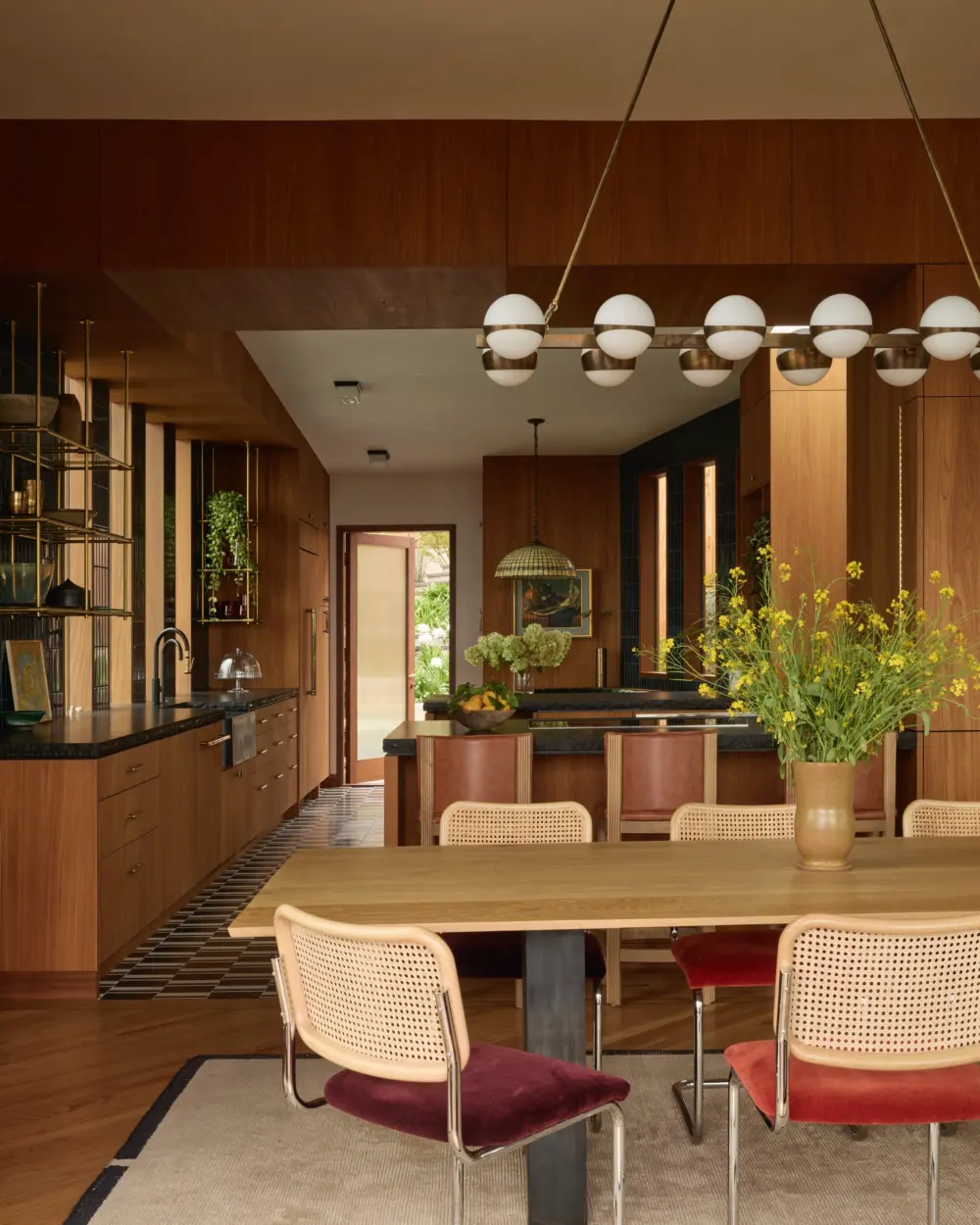Interior: kitchen-dining room with walnut cabinetry, black countertops, mid-century dining set with cane-back chairs, globe lighting, garden view.
