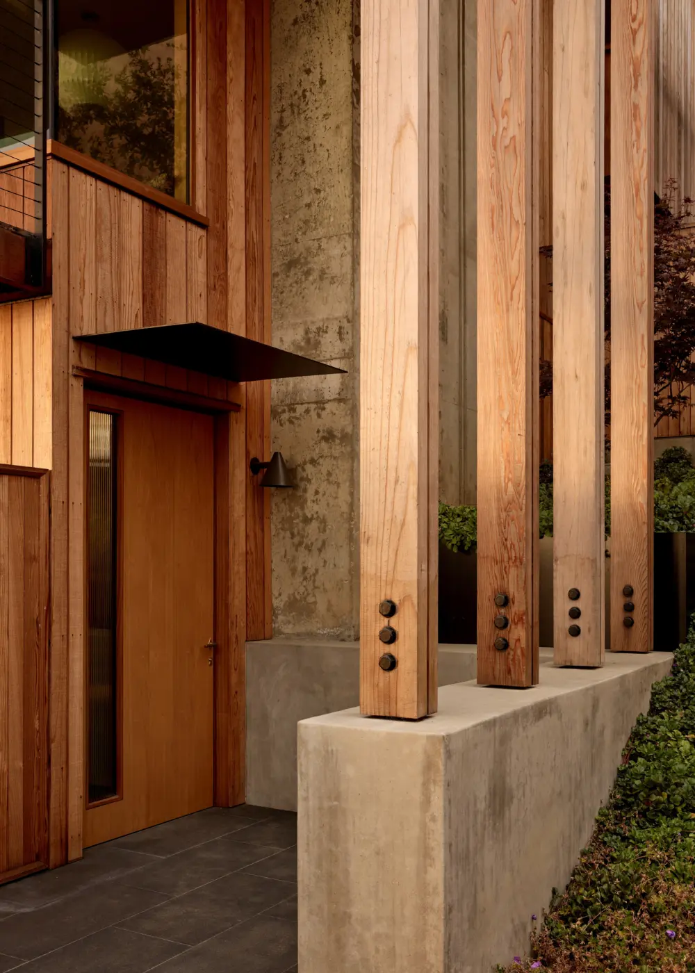 Exterior entry with tall timber columns on a concrete base, wood-clad facade, and a black metal canopy over a wooden door; hillside planting.