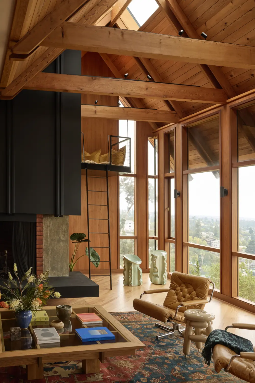 Timber-framed great room with floor-to-ceiling windows, exposed trusses, brick fireplace, mezzanine ladder; leather lounge chairs, glass coffee table, city view.