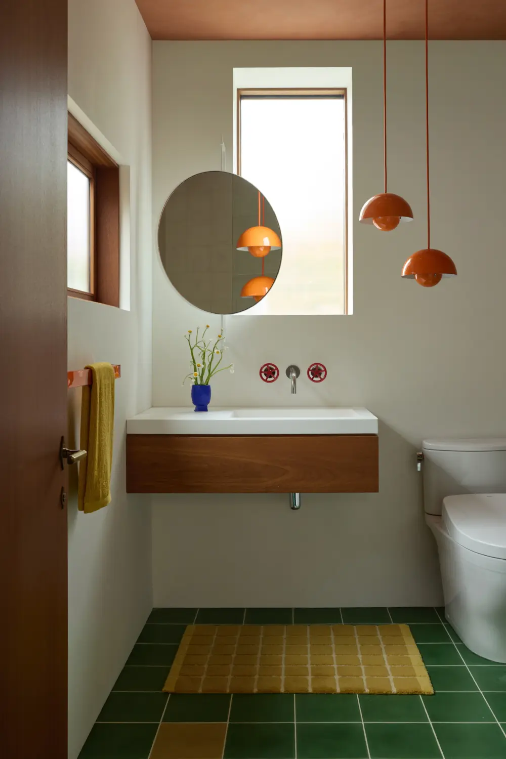 Interior bathroom with wall-mounted walnut vanity, round mirror, orange pendant lights, white sink, green tile floor, yellow rug, and toilet.