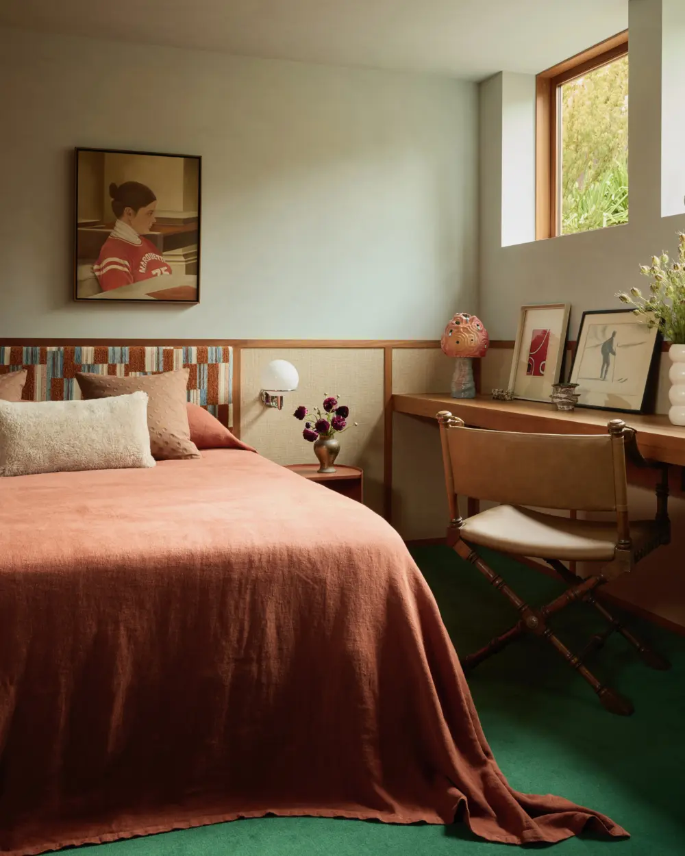 Interior bedroom in a custom-home: queen bed with coral quilt, woven headboard, wood desk by high windows, framed art, green carpet.