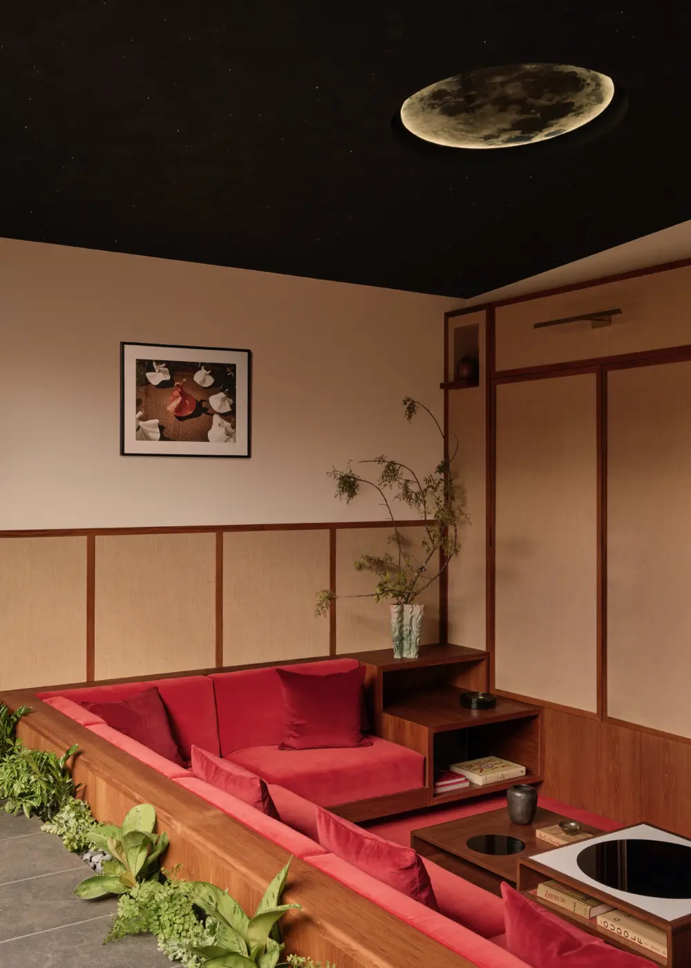 Interior living alcove with L-shaped built-in wooden bench, red velvet cushions, woven wood wall panels, potted plant, low coffee table, moon-shaped ceiling light.