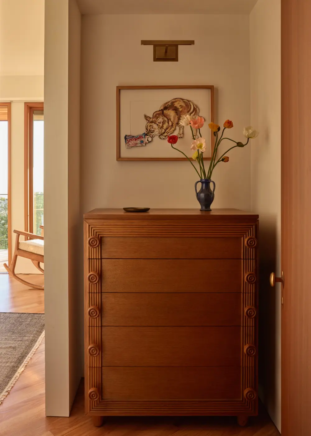 Interior entry alcove: oak dresser with carved vertical columns, blue vase with flowers, framed cat artwork above; adjacent room with large windows.