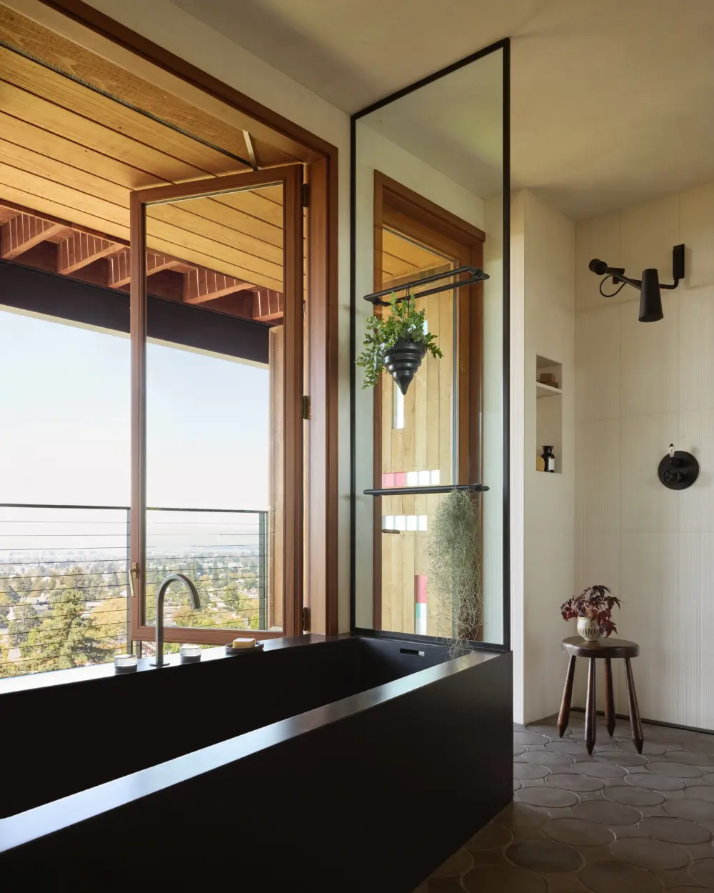 Interior bathroom with black rectangular soaking tub by wood-framed windows, glass divider, white tile wall, hanging plant, and hillside city view.