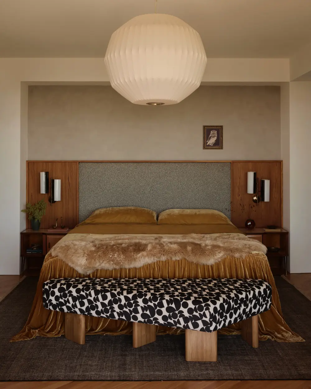 Bedroom interior with walnut headboard, gray panel, mustard velvet bedspread, fur throw, black-and-white patterned bench, and white pendant light.