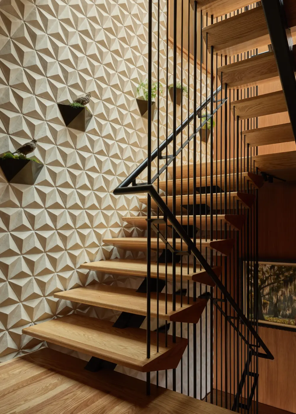 Interior staircase with open oak treads and black metal balusters beside a 3D geometric wall with small planters and greenery.
